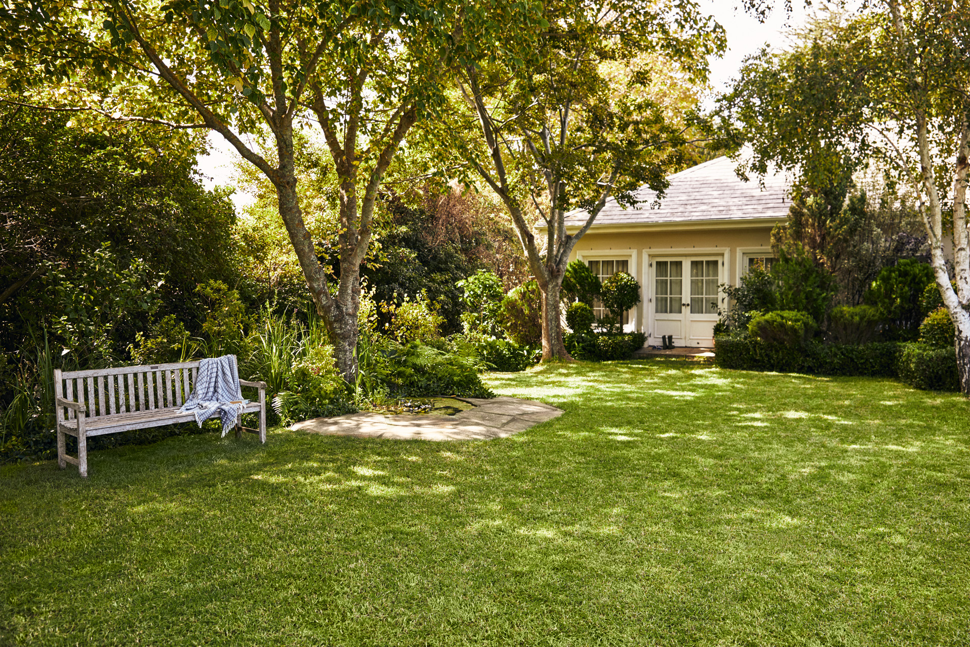 Malý domek se zahradou, v popředí zahradní lavička na zeleném trávníku